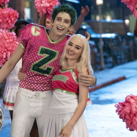 In the foreground, green-haired zombie Zed (Milo Manheim) wears a pink and green Seabrook football jersey. He has removed his helmet and is hugging blonde cheerleader Addison (Meg Donnelly), who is wearing a pink, green, and white uniform. In the background, Seabrook cheerleaders are waving pink pom poms in the air.