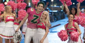 In the foreground, green-haired zombie Zed (Milo Manheim) wears a pink and green Seabrook football jersey. He has removed his helmet and is hugging blonde cheerleader Addison (Meg Donnelly), who is wearing a pink, green, and white uniform. In the background, Seabrook cheerleaders are waving pink pom poms in the air.