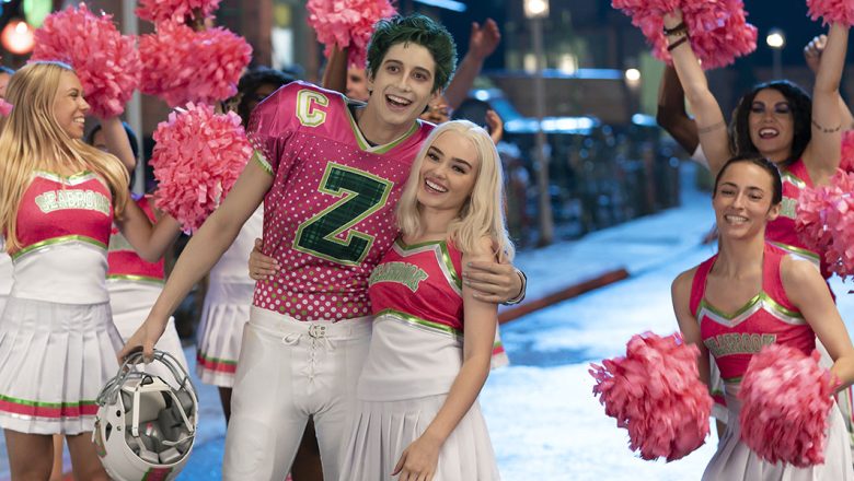 In the foreground, green-haired zombie Zed (Milo Manheim) wears a pink and green Seabrook football jersey. He has removed his helmet and is hugging blonde cheerleader Addison (Meg Donnelly), who is wearing a pink, green, and white uniform. In the background, Seabrook cheerleaders are waving pink pom poms in the air.