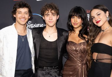 Matt Cornett, Joshua Bassett, Sofia Wylie, and Olivia Rodrigo wrap their arms around each other on the red carpet at the Season 3 premiere of High School Musical: The Musical: The Series. Matt is wearing an embroidered white jacket over a slate blue shirt and navy blue dress pants. He accessorized his ensemble with a silver cross. Joshua is wearing a black satin suit and a black tank top, which he accessorized with a pendant necklace. Sofia is wearing a shimmering, strapless brown mini dress and hoop earrings. Olivia is wearing a black, midriff-baring dress that features a sheer panel of fabric on her left side. She accessorized with a chain necklace and silver earrings. They are standing in front of a black backdrop featuring a red and white High School Musical: The Musical: The Series logo and a white Disney+ logo.