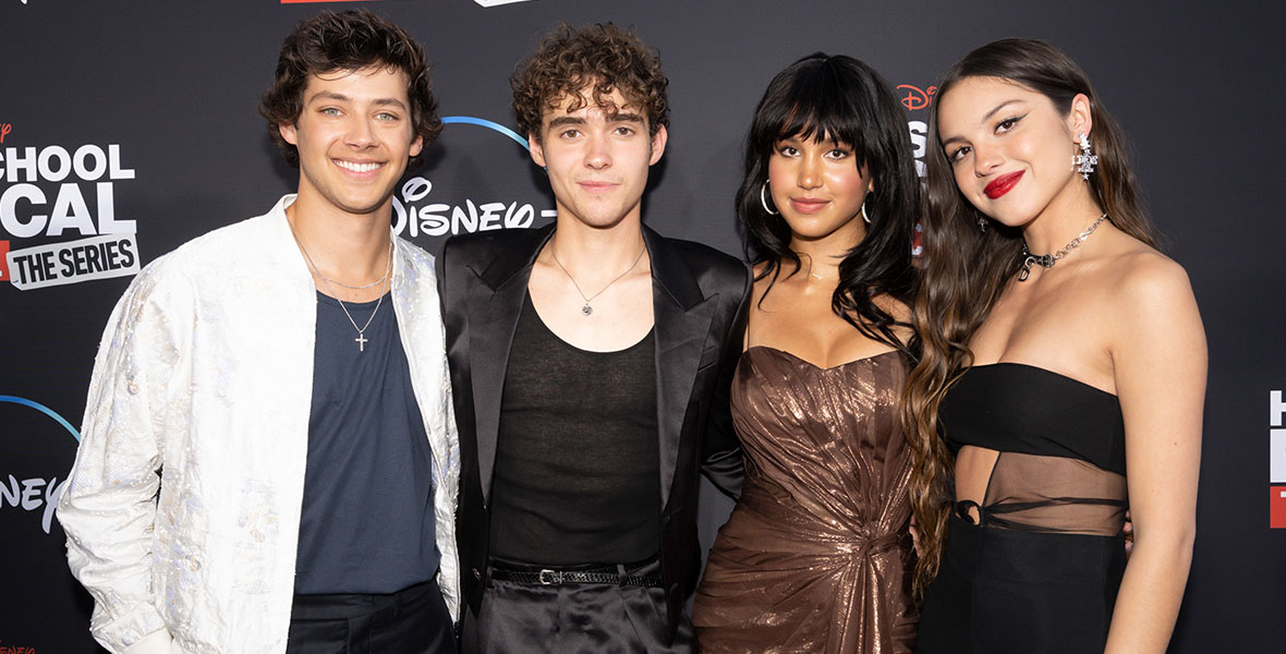 Matt Cornett, Joshua Bassett, Sofia Wylie, and Olivia Rodrigo wrap their arms around each other on the red carpet at the Season 3 premiere of High School Musical: The Musical: The Series. Matt is wearing an embroidered white jacket over a slate blue shirt and navy blue dress pants. He accessorized his ensemble with a silver cross. Joshua is wearing a black satin suit and a black tank top, which he accessorized with a pendant necklace. Sofia is wearing a shimmering, strapless brown mini dress and hoop earrings. Olivia is wearing a black, midriff-baring dress that features a sheer panel of fabric on her left side. She accessorized with a chain necklace and silver earrings. They are standing in front of a black backdrop featuring a red and white High School Musical: The Musical: The Series logo and a white Disney+ logo.