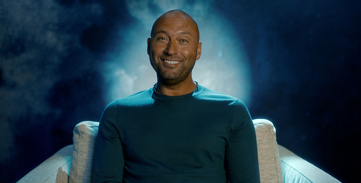 Former MLB player Derek Jeter smiles as he sits in a gray chair in front of a blue backdrop in ESPN Films’ The Captain.
