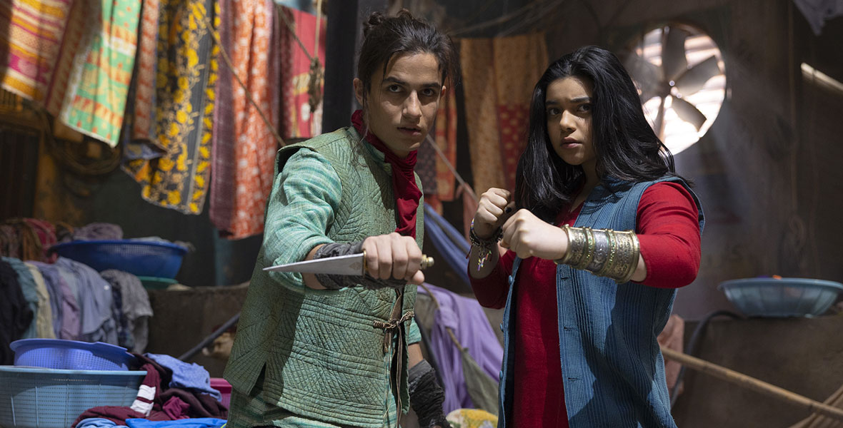 (L-R): In Marvel Studios’ Ms. Marvel, Aramis Knight as Red Dagger/Kareem and Iman Vellani as Ms. Marvel/Kamala Khan stand with their hands ready to attack. Aramis is wearing a green vest and a red scarf and wields a dagger, while Kamala wears a blue vest over a red tunic and a mystical bangle. They are surrounded by various colorful linens.