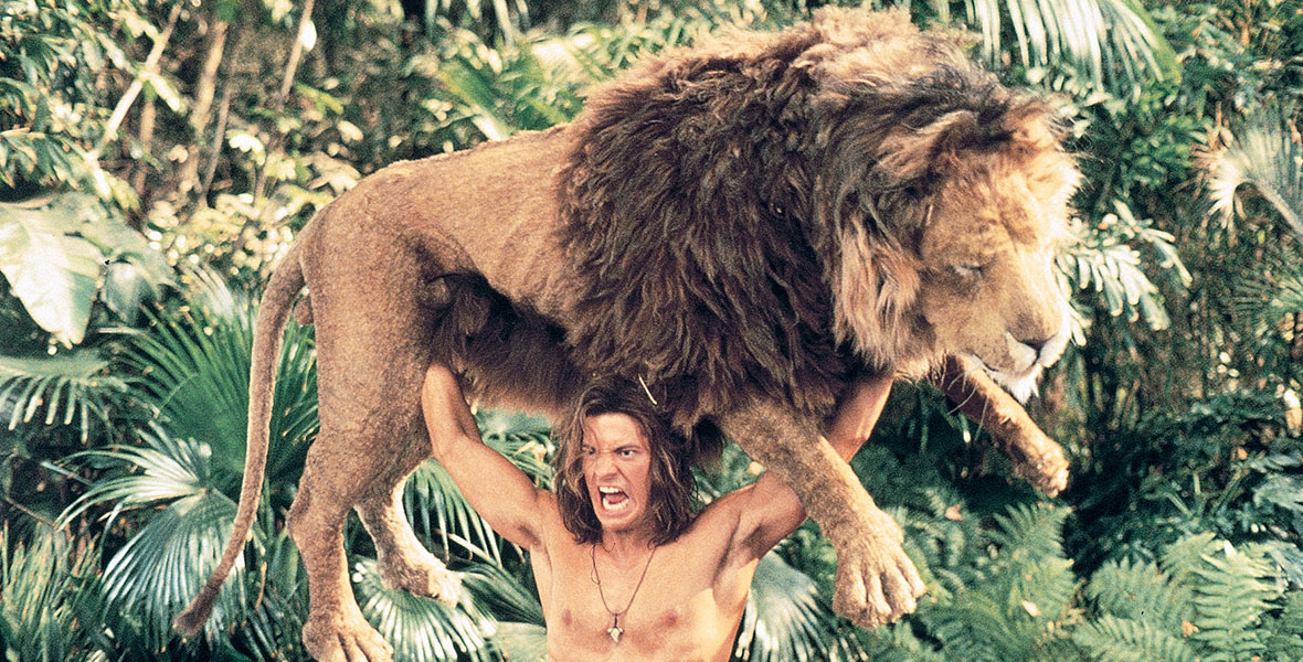 Actor Brendan Fraser, shirtless, holds a large lion above his head while standing in the African jungle.