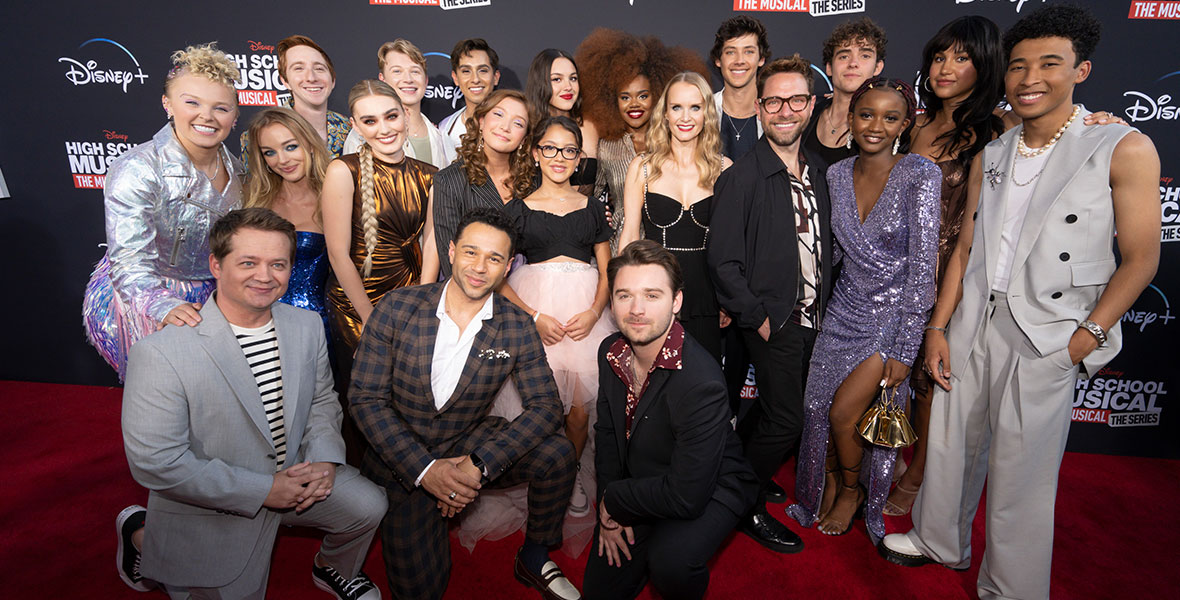 The cast and creator/executive producer of High School Musical: The Musical: The Series pose for a group photo on the Season 3 premiere red carpet. Back row, left to right: Larry Saperstein, Joe Serafini, Frankie Rodriguez, Olivia Rodrigo, Dara Reneé, Matt Cornett, Joshua Bassett, Sofia Wylie, and Adrian Lyles. Middle row, left to right: JoJo Siwa, Olivia Rose Keegan, Meg Donnelly, Saylor Bell Curda, Liamani Segura, Kate Reinders, Tim Federle, Aria Brooks. Front row, left to right: Jason Earles, Corbin Bleu, Ben Stillwell. They are posing in front of a black backdrop featuring a red and white High School Musical: The Musical: The Series logo and a white Disney+ logo.