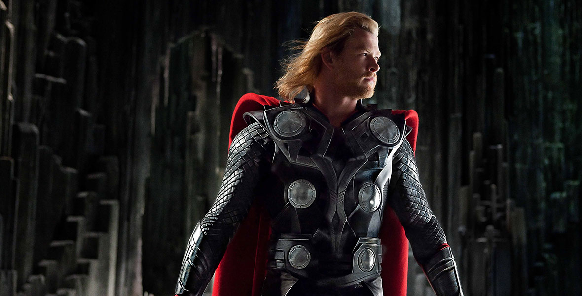 Chris Hemsworth, as Thor, stands in armor and a red cape looking to his left as he holds his hammer in his right hand in front of a dimly lit background.