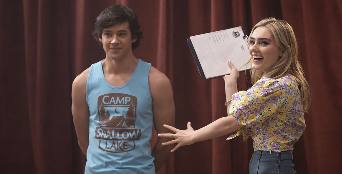 Matt Cornett (left) and Meg Donnelly (right) in front of a red show curtain. Cornett is standing with his hands behind his back and is wearing a light blue tank top that has a Camp Shallow Lake logo on it in brown. Donnelly is gesturing toward Cornett with a big smile on her face. She has a clipboard in her right hand, as if she is organizing something for their performance. She is wearing high-waisted jeans and a purple and yellow floral print top.