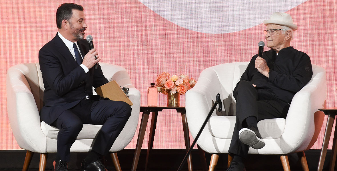 Jimmy Kimmel, on left, and Norman Lear, on right, are seated in white chairs and talking into microphones, in front of a pink-hued background. Jimmy is wearing a black suit and tie, and Norman is wearing a black sweater and pants and a white fedora, and a cane is resting next to him against the chair.There is a table between them, on which sits a vase of pink flowers.