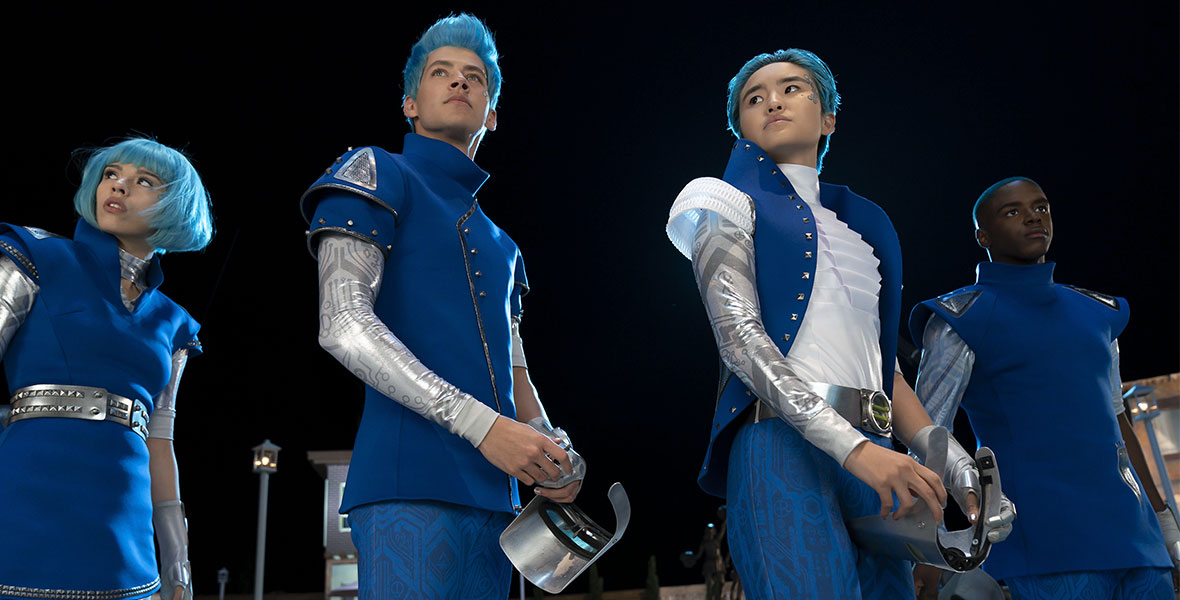 (Left to Right): Aliens A-li (Kyra Tantao), A-lan (Matt Cornett), and A-Spen (Terry Hu) stand in the town square of Seabrook at night. They have icy blue hair and are wearing futuristic outfits in shades of blue, silver, and white.