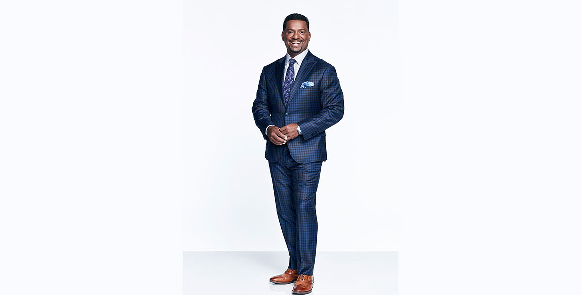 New Dancing with the Stars co-host Alfonso Ribeiro is wearing a blue checkered suit and brown shoes, standing against a white background, and is smiling at the camera.