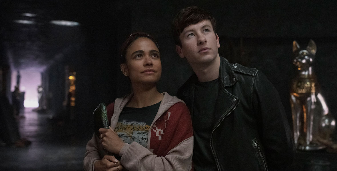 Inside The Domo, the starship used by the Eternals, Makkari, played by Lauren Ridloff, leans against Druig, played by Barry Keoghan, as they both look up. Makkari is holding an ancient artifact and wearing a graphic tee, a knit hoodie, and delicate jewelry. Druig is wearing a black leather jacket and a black shirt.