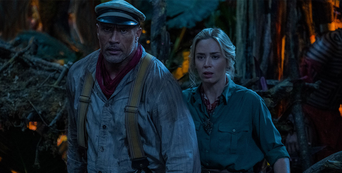 Actor Dwayne Johnson and Emily Blunt lock arms while passing through a dark jungle at nighttime in a still from Jungle Cruise.