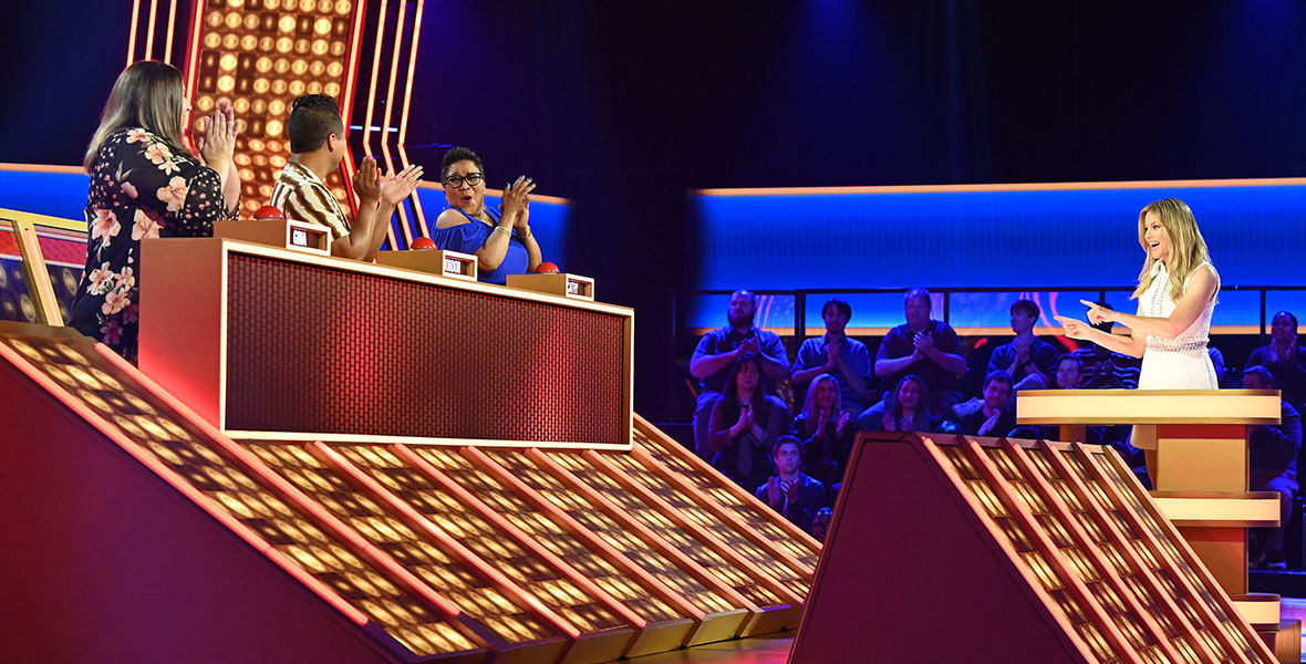 Host Elizabeth Banks (right) points to a trio of contestants on the ABC game show Press Your Luck. Elizabeth is wearing a white dress and standing at a podium. To her right, the three contestants—Gina, Jordon, and Cathy—are seated next to each other and clapping.