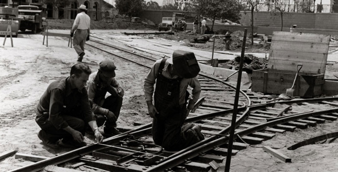 Three men assemble railroad tracks at Disneyland Park with additional construction work occurring in the background.