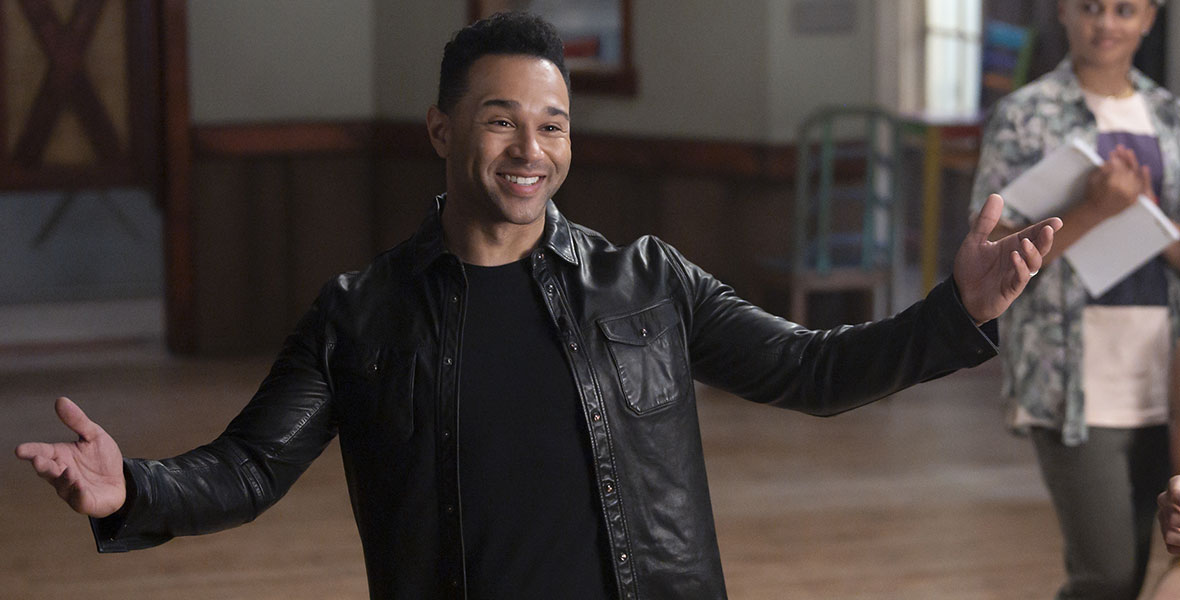 Corbin Bleu stands in a black t-shirt, black pants and a black leather jeacket with his arms stretched out on either of his sides. He is grinning and has short dark hair.