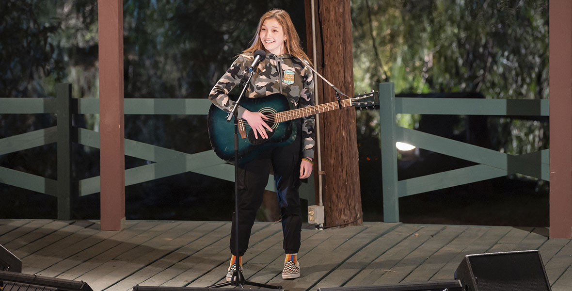 In a still from season three of High School Musical: The Musical: The Series, Saylor Bell Curda as Maddox is standing on a wooden stage, with a banner above her reading “Camp Shallow Lake.” She is holding a guitar and standing at a microphone, smiling, and she’s wearing a camo sweatshirt and black pants.