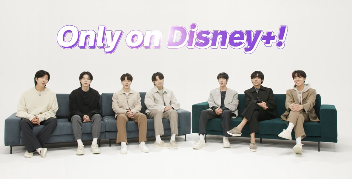 The members of BTS, dressed in various shades of white, black, tan, and gray, are sitting split up between two couches—one grey, one green—set against a white background. Above them, in white and purple writing, it says “Only on Disney+!”