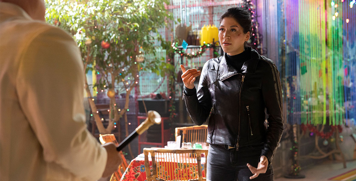 Maya Lopez, played by Alaqua Cox, uses American Sign Language (ASL) to communicate with Kingpin (Vincent D’Onofrio), in a scene from Marvel Studios’ Hawkeye. Maya is wearing a black leather jacket and skintight jeans, and her hair is pulled back. She has cuts and bruises on her face. Kingpin, whose back is turned to the camera, is holding a cane and wearing a white blazer. They are standing inside a colorful restaurant that is decorated for Christmas with garland, ornaments, and more.