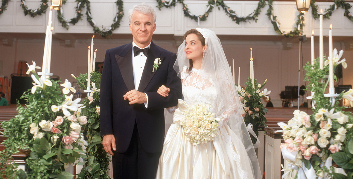 A man, played by Disney Legend Steve Martin, walks his daughter, played by Kimberly Williams, down the aisle. He is wearing a classic tuxedo and she is wearing a lacy wedding gown and a delicate veil. She is carrying white flowers. The church pews are lined with white and pink roses and lilies.