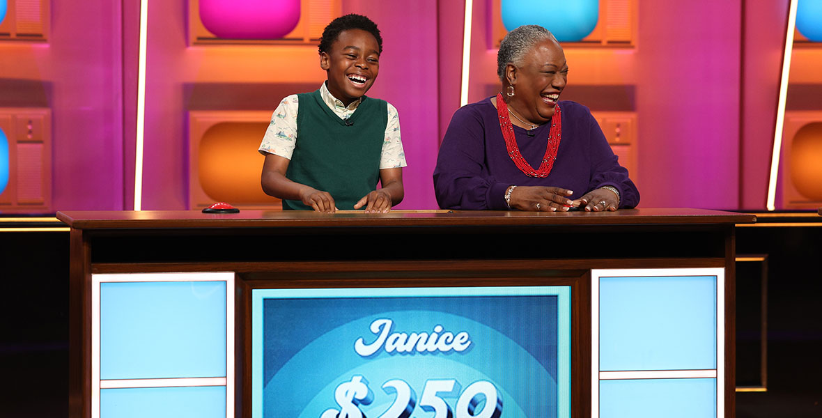 Two contestants from the ABC quiz show Generation Gap await their next question from behind a brown podium with a mounted screen. A young boy, wearing a green vest, is laughing as he sits beside his elder, Janice, who is laughing and wearing a purple top and a red beaded necklace.