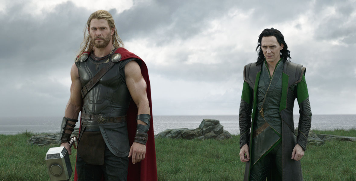(L-R) Chris Hemsworth as Thor and Tom Hiddleston as Loki stand in a lush grassy field in Marvel Studios’ Thor: Ragnarok.