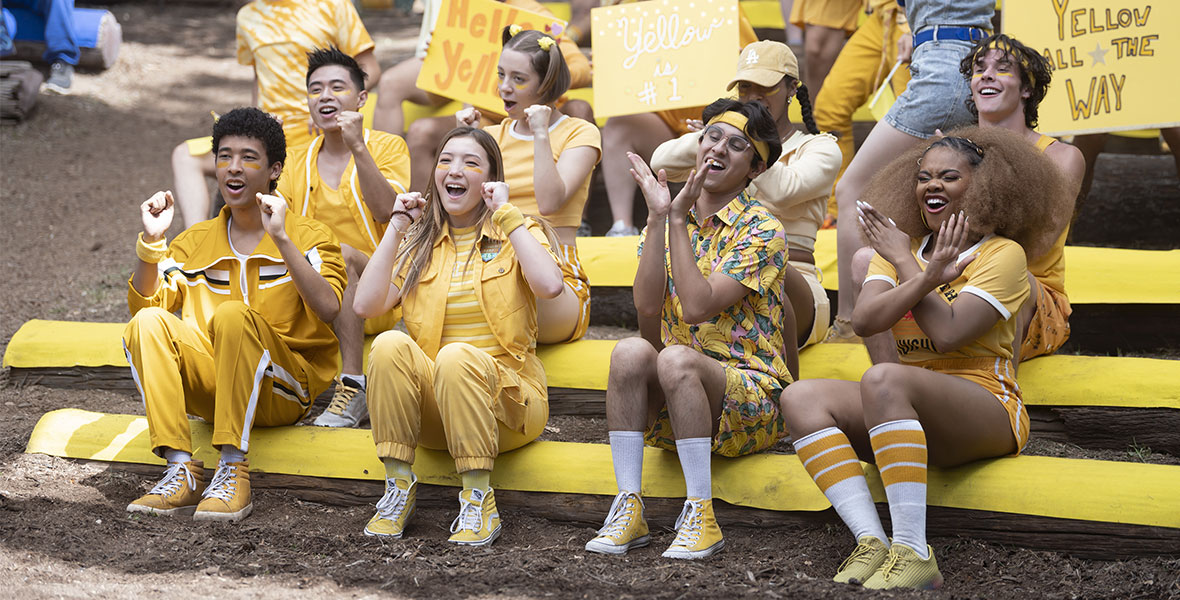 In a still from season three of High School Musical: The Musical: The Series, from left to right, Adrian Lyles as Jet, Saylor Bell Curda as Maddox, Frankie Rodriguez as Carlos, and Dara Reneé as Kourtney are all wearing bright yellow outfits and participating in a “color war” pep rally at Camp Shallow Lake, surrounded by fellow campers sitting on rows of logs.