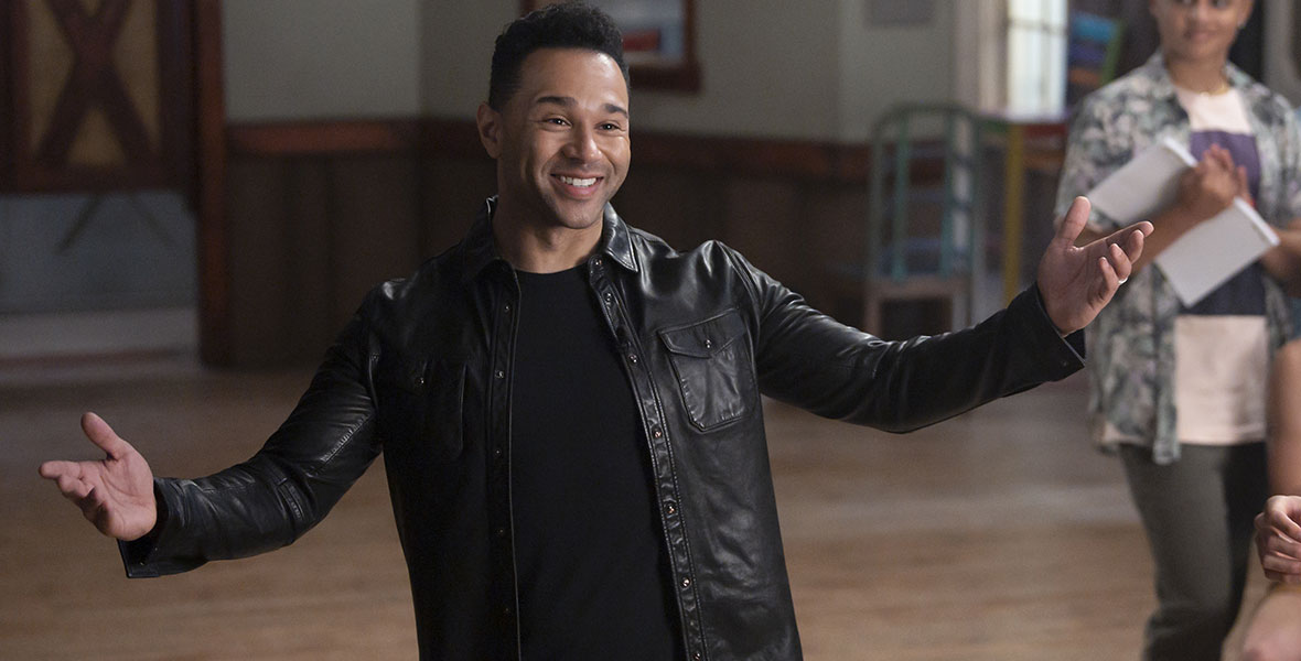 Corbin Bleu stands confidently with his arms outstretched and a huge smile on his face. His black hair is short and faded. He has on black pants, a black shirt, and a black leather jacket. He seems to be standing in a camp recreation center, with one camper blurry, but visible behind him.