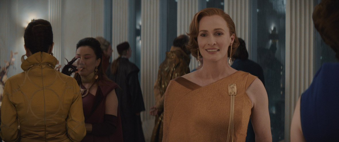 Genevieve O’Reilly as Mon Mothma. She is in a party setting with people dressed nicely around her eating, drinking, and talking. Mon Mothma is looking just left of camera. She has short red hair and is wearing an asymmetrical gold dress with a large gold broch on her left and long gold drop earrings.