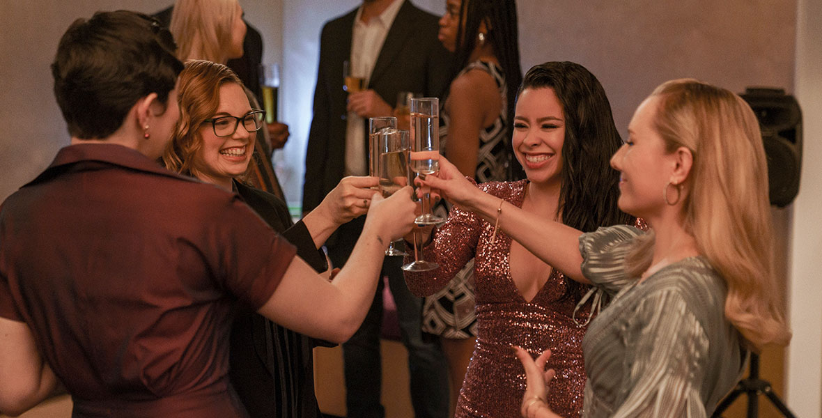 The FCG crew stand in a circle with the champagne glasses in the air as they make a toast during an episode of Good Trouble. Mariana, played by actress Cierra Ramirez, stands in the center of a group wearing a sequined dress with a plunging neckline.