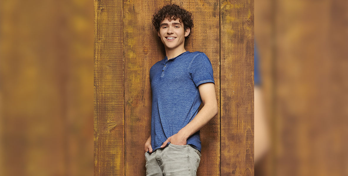 Curly brown-haired Joshua Bassett leans with his right shoulder against a dark wooden wall. He is wearing a dark blue T-shirt and gray pants. Both of his hands are tucked into his pockets.