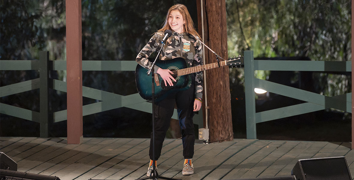 Saylor Bell as Maddox in a still from High School Musical: The Musical: The Series Season 3. Maddox is standing alone on an outdoor stage under a sign that reads “Camp Shallow Lake.” She is smiling out at the audience and casually holding her blue acoustic guitar, which is hanging around her shoulders. She is wearing a gray and pink camouflage pattern hoodie, navy blue jogger pants, and slip-on shoes with rainbow socks. She looks as if she just finished a performance.