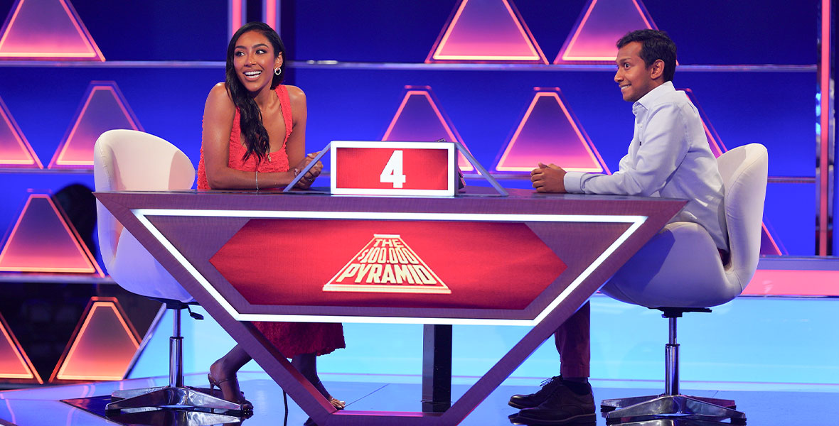 Former Bachelorette star Tayshia Adams sits at a table across from a contestant on The$100,000 Pyramid, awaiting instructions from host Michael Strahan.