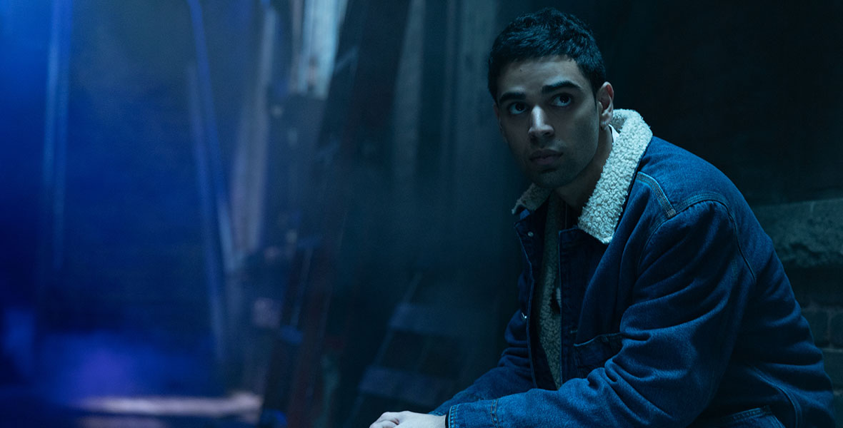 Actor Rish Shah sits in a dimly lit room wearing a jean jacket and slacks in Marvel Studios’ Ms. Marvel.