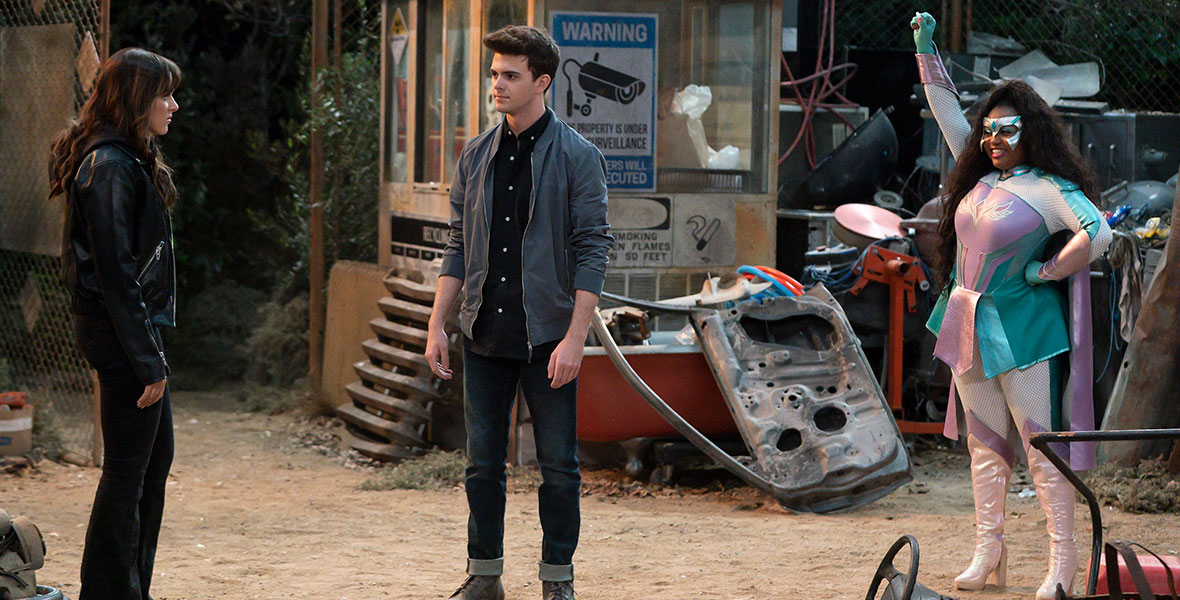 Isabella Pappas (left), Reed Horstmann (center), and Mariah Iman Wilson (right) stand in a junkyard as Wilson dons a brightly colored super hero ensemble.