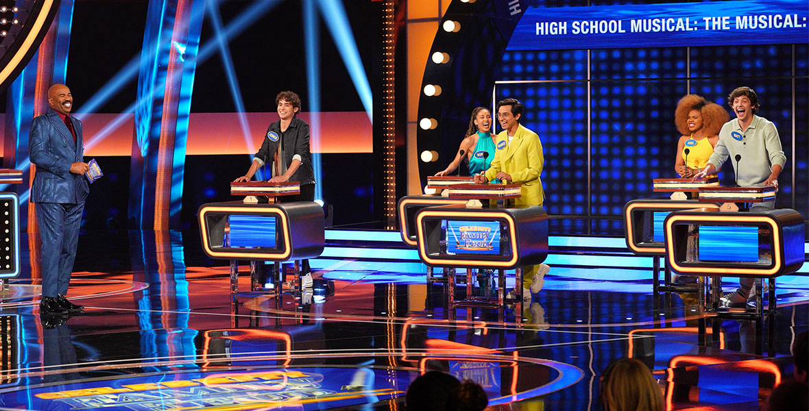 (Left to right) Host Steve Harvey stands next to actors Joshua Bassett, Sofia Wylie, Frankie A. Rodriguez, Dara Renee, and Matt Cornett, who each have solo podiums and are surrounded by bright stage lights and graphics.