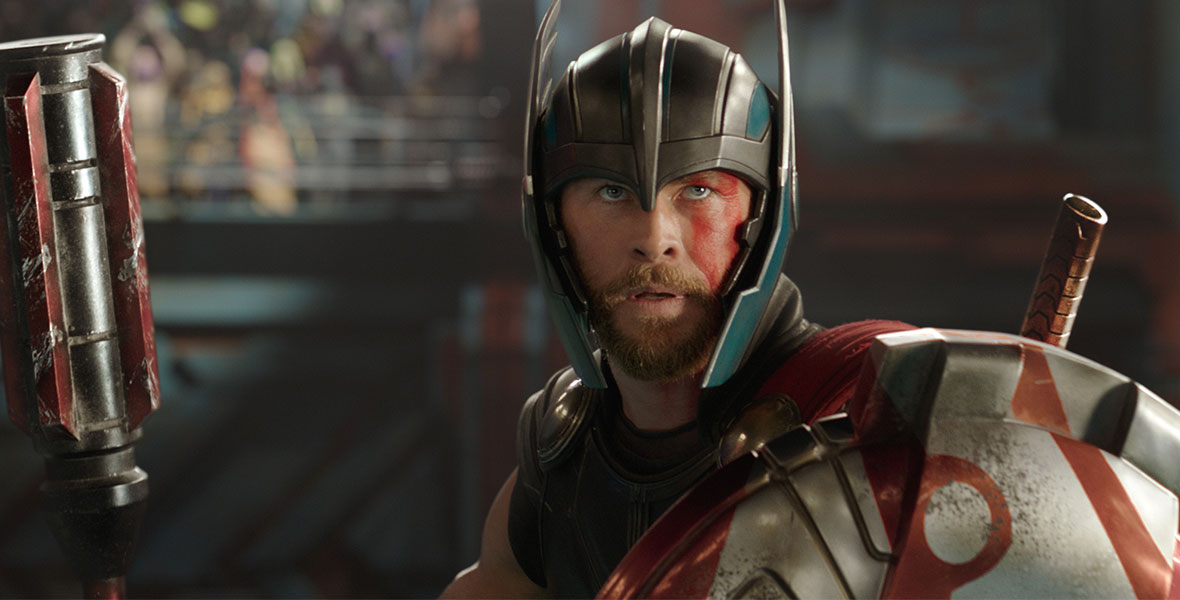 In a medium close-up, Thor stands in a metal helmet and large metal amor over his shoulders.