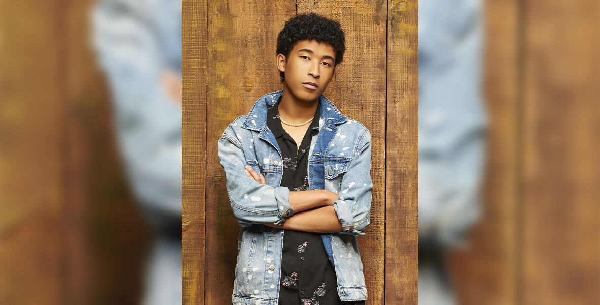 A headshot image of Adrian Lyles (Jet). Lyles is standing against a wooden wall with his arms crossed in front of him. He isn’t smiling and has a serious expression on his face. He has short, dark, curly hair and is wearing a bleach-spotted denim jacket over a black Hawaiian shirt with palm trees and beach shacks on it.