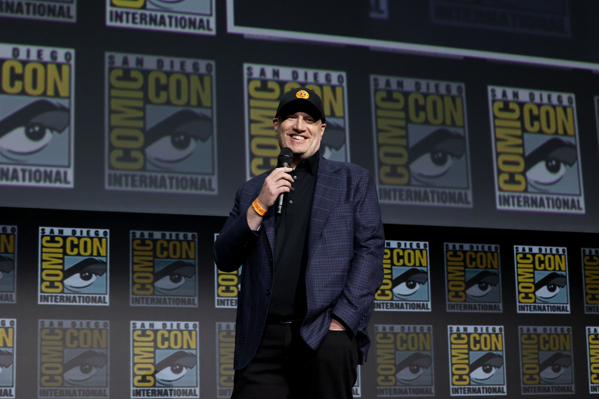Kevin Feige onstage at San Diego Comic-Con. He is wearing a blue checkered suit jacket over a black polo shirt and black pants. He is wearing a black baseball cap with Miss Minutes from Loki on it. He is holding a mic and behind him is a screen with the San Diego Comic-Con logo repeating.