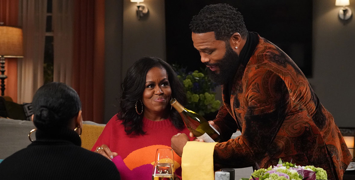 Michelle Obama smiles as Dre, played by Anthony Anderson, pours her a glass of white wine. She is seated opposite Bow, played by Tracee Ellis Ross, at the dinner table. On the table, there is a plate of salad, a stick of butter, salt and pepper shakers, and a floral arrangement.