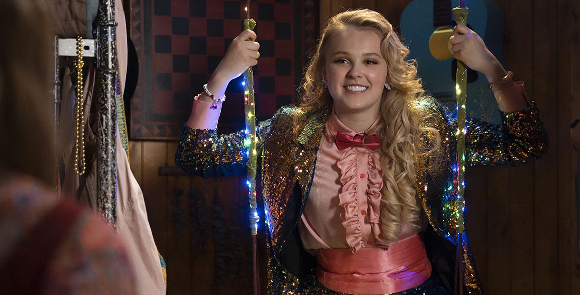 Jojo Siwa as Madison in High School Musical: The Musical: The Series. She is sitting on a wooden swing that has colorful lights wrapped around the side ropes. She is wearing a gold, bedazzled pantsuit, a pink ruffled dress shirt underneath, with hot pink bowtie and cummerbund. Jojo’s long blonde hair is style in loose curls and she is smiling at someone off-camera and grabbing the sides of the swing with both hands.