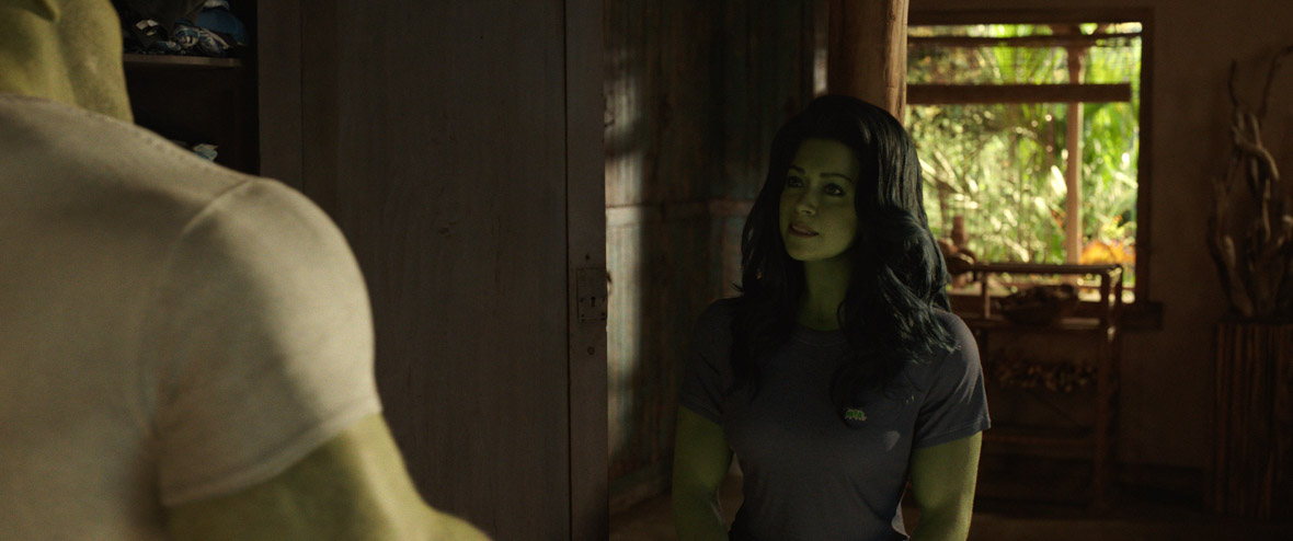 Tatiana Maslany as She-Hulk in a screen shot from the show. On the left you can see The Hulk facing away from camera; you can see the back of his green head and his green arm and he is wearing a white T-shirt. Maslany is wearing a gray T-shirt and her hair is down in loose waves.