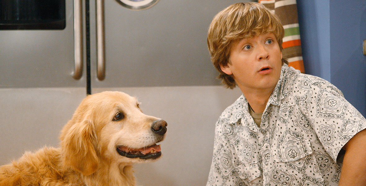 Jason Earles is squatting next to a golden retriever on his right. He is wearing a white and grey paisley patterned button-up short sleeve shirt with a muted khaki underneath. His hari is light brown and his eyes are looking to his left in a glimpse of shock.