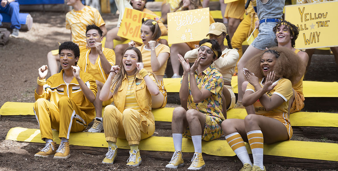 A group of campers during a “Color War” scene from High School Musical: The Musical: The Series. There are multiple rows of campers sitting on yellow beams and they are all wearing yellow. In the front row, from left to right, you see Adrian Lyles, Saylor Bell, Frankie Rodriguez, and Dara Reneé. They are cheering, clapping, and singing. Campers behind them are holding signs that say “Hello Yellow,” “Yellow is #1”, and “Yellow All the Way”. Meg Donnelly is on the right side of the image in a denim jumpsuit and holding a mic.