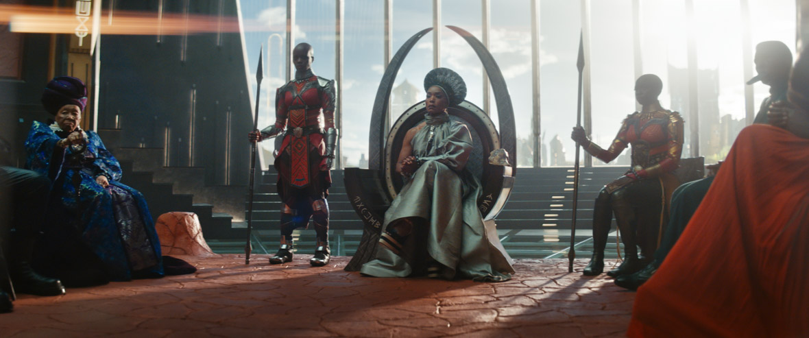 A screenshot from Black Panther: Wakanda Forever. Angela Bassett as Ramonda sits in the main throne with two Dora Milage on either side of her. Her throne is circular and has two large talon shaped pieces arching above her head. Wakandan writing can be seen on the seat of the throne. The Dora Milage are in their classic red and black armor and both have tall spears in hand. There is another council member to the queen’s left. She is very old and wearing deep blue robes. Behind the throne room is a wide window showing the daylight sky.