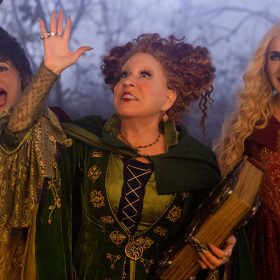 Still from Disney’s Hocus Pocus 2, featuring (from left to right) Kathy Najimy as Mary Sanderson, Disney Legend Bette Midler as Winifred Sanderson, and Sarah Jessica Parker as Sarah Sanderson. Winifred is wearing her signature green witch’s dress and cloak and is holding her book of spells, and her arm is up in the air as if conjuring something. All three are looking excitedly up at the sky, with spooky trees in the background.