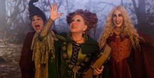 Still from Disney’s Hocus Pocus 2, featuring (from left to right) Kathy Najimy as Mary Sanderson, Disney Legend Bette Midler as Winifred Sanderson, and Sarah Jessica Parker as Sarah Sanderson. Winifred is wearing her signature green witch’s dress and cloak and is holding her book of spells, and her arm is up in the air as if conjuring something. All three are looking excitedly up at the sky, with spooky trees in the background.