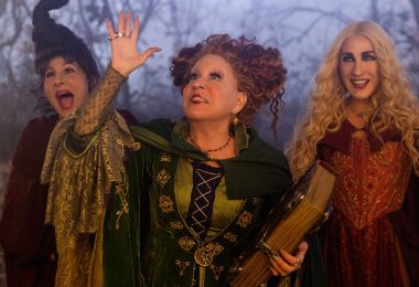 Still from Disney’s Hocus Pocus 2, featuring (from left to right) Kathy Najimy as Mary Sanderson, Disney Legend Bette Midler as Winifred Sanderson, and Sarah Jessica Parker as Sarah Sanderson. Winifred is wearing her signature green witch’s dress and cloak and is holding her book of spells, and her arm is up in the air as if conjuring something. All three are looking excitedly up at the sky, with spooky trees in the background.