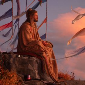In a production still from Marvel Studios’ Thor: Love and Thunder, actor Chris Hemsworth, as Thor, meditates while sitting under a flowering tree. Blue and red flags hang from the tree’s branches, and the clouds in the sky behind him have a pink hue.