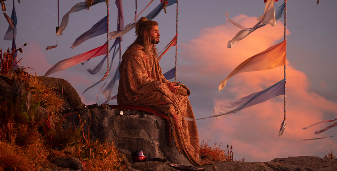 In a production still from Marvel Studios’ Thor: Love and Thunder, actor Chris Hemsworth, as Thor, meditates while sitting under a flowering tree. Blue and red flags hang from the tree’s branches, and the clouds in the sky behind him have a pink hue.