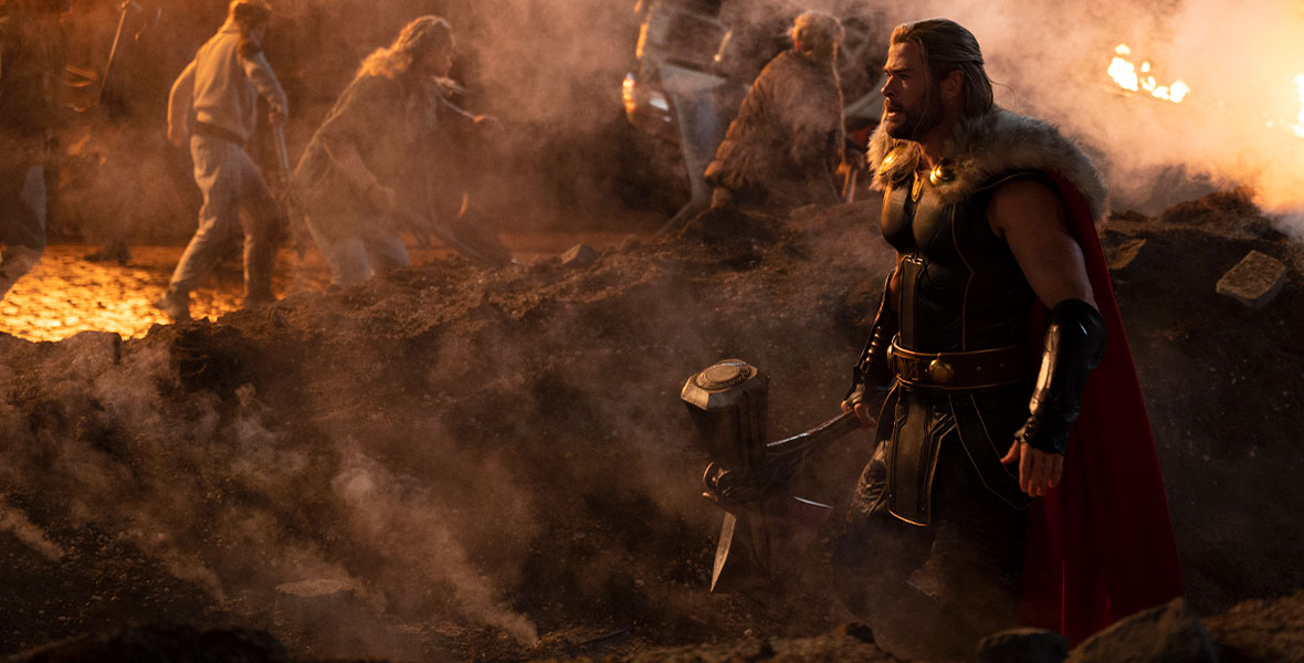 In a production still from Marvel Studios’ Thor: Love and Thunder, actor Chris Hemsworth, as Thor, stands in a cloud of smoke. In the background, the people of New Asgard fight a mysterious enemy force.